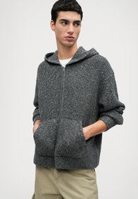 Mladý muž v šedej pletenej mikine na zips s prednými vreckami a béžovými nohavicami, stojí s rukami vo vreckách, pozerá sa do strany.