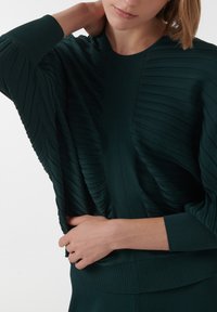 Donkergroen geribbelde top met gestructureerde strepen, een brede halslijn en lange mouwen. Stof lijkt zacht met een aansluitende zoom.