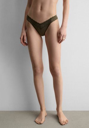 Frau steht barfuß und trägt einen olivgrünen Bikini-Slip mit Spitzenbesatz vor einem schlichten grauen Hintergrund.