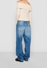 Frau in einem ärmellosen beige Top, lockeren, ausgewaschenen blauen Jeans mit Gesäßtaschen und schwarzen klobigen Schuhen, die mit dem Rücken zur Kamera steht.