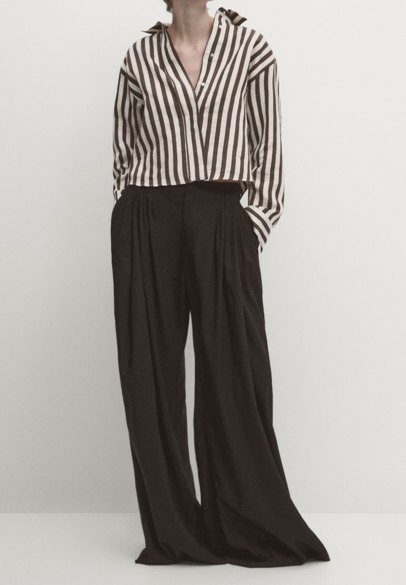 Personne portant une chemise courte à manches longues rayée verticalement marron et blanche, avec un pantalon noir plissé à jambes larges, les mains dans les poches.