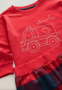 Red long-sleeve shirt with white embroidered ambulance and "whoosh" text; features ribbed neckline and cuffs, paired with a plaid skirt.