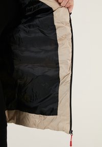 Insulated jacket with a beige outer shell and black lining; features a front zipper and red pull tab. Soft texture with a smooth finish.