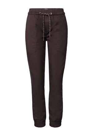 Gros plan d'une personne portant un pantalon noir à cordon et un haut en maille beige, tenant un sac bandoulière beige tissé sur le côté gauche.