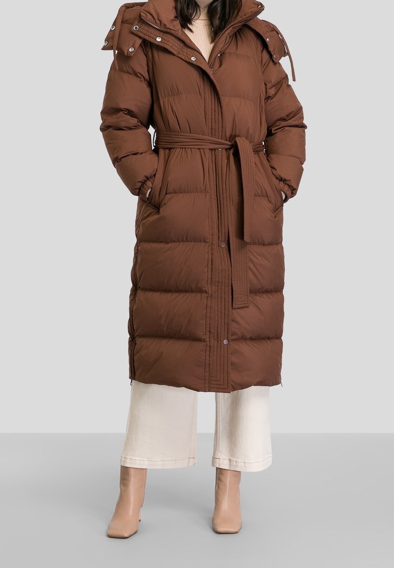 Kvinna klädd i en lång brun vadderad jacka med bälte, vita vida byxor och beige ankelskor, stående mot en grå bakgrund.