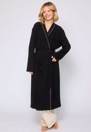 Robe noire à manches longues avec des bordures blanches, dotée de deux poches à l'avant et d'une ceinture assortie. Portée par-dessus un pantalon de couleur claire.