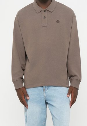 Mand iført en brun polotrøje med knapper og lyseblå jeans, stående foran en ensfarvet baggrund.