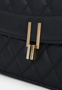 Zbliżenie na pikowaną czarną torebkę skórzaną, z pozłacaną metalową klamrą i teksturowanymi akcentami. Powierzchnia ma miękkie wykończenie z ukośnym przeszyciem.