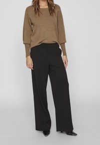 Pull en tricot marron à manches longues associé à un pantalon noir ample et des chaussures noires à bouts pointus. Design simple, texture douce, sans motifs visibles.