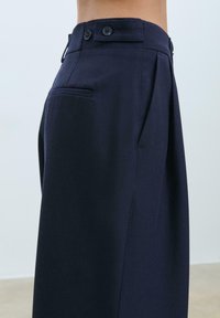 Pantaloni blu navy in tessuto morbido, con design plissettato, due bottoni in vita e una tasca laterale.