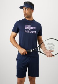 Άνδρας σε ναυτικό αθλητικό ένδυμα και καπέλο, κρατώντας ένα ρακέτα του τένις, κοιτάζοντας δεξιά σε έναν απλό φωτεινό φόντο.