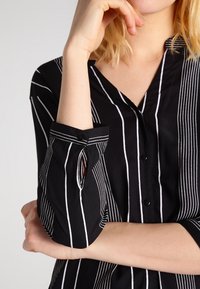 Femme portant un chemisier noir à rayures blanches verticales, un bras plié avec la main près du visage et l'autre bras croisé sur la taille.