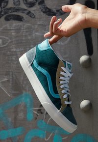 Tenisky vysokého střihu s teplými a béžovými kordurovými panely, bílými tkaničkami a bílou gumovou podrážkou, zavěšené proti graffiti pozadí.