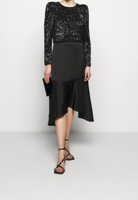 Kobieta w czarnej, cekinowej bluzce z długim rękawem, asymetrycznej czarnej midi spódnicy, czarnych, zdobnych szpilkach oraz trzymająca czarną kopertówkę.