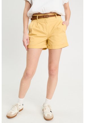 Pantaloni corti gialli in cotone con una cintura intrecciata marrone, dotati di tasche laterali e orlo arrotolato. Indossati con una camicia bianca e sneakers.