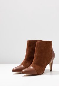 Botas de tobillo de ante marrón con punta afilada y una tapa de cuero marrón en contraste. Presentan un tacón de aguja y una textura suave.