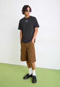 Czarny T-shirt z krótkim rękawem z logo, brązowe szorty cargo z kieszeniami, białe skarpetki, czarne loafersy z frędzlami na zielonej podłodze.