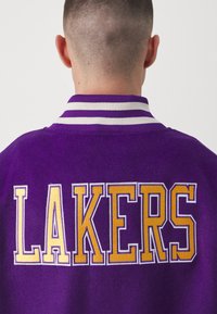 Chaqueta morada con un destacado bordado de "LAKERS" en amarillo. El cuello con rayas blancas aporta contraste al material de felpa texturizada.