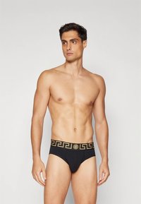 Άνδρας χωρίς μπλούζα που φορά μαύρα σλιπ με ζώνη με σχέδιο χρυσού ελληνικού κλειδιού και έμβλημα της Μέδουσας, στέκεται σε απλό φόντο.