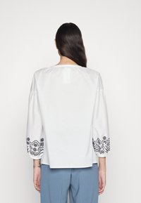 Blouse en coton blanc avec des manches bouffantes trois-quarts. Une broderie florale noire orne les poignets, avec des motifs intriqués le long de l'ourlet.