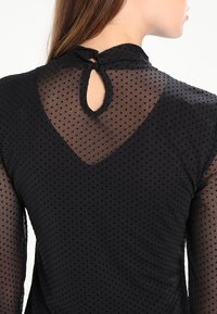 Schwarze gepunktete Netzbluse mit langen Ärmeln, einer Schlüssel-Ausschnitt-Rückenschließe und strukturiertem Stoff mit kleinen, gleichmäßig verteilten Pünktchen.