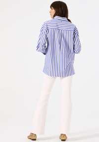Camisa de botón a rayas azul y blanca con un corte holgado, combinada con pantalones blancos acampanados y zapatos con acentos dorados.