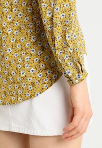 Blouse jaune à manches longues avec un motif floral blanc et bleu portée avec une jupe blanche, montrant le détail du poignet et la main reposant sur le côté.