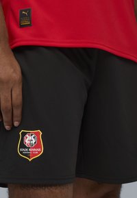Camiseta deportiva roja de tejido que absorbe la humedad y pantalones cortos negros con un logo bordado del Stade Rennais en rojo, amarillo y blanco.