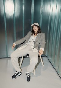 En person i en grå rutiga kavaj över en krämfärgad tröja, beige byxor och svarta sneakers, sitter på en ljus trästol mot en metallisk bakgrund.