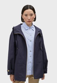 Navy blue hooded jacket with a zip front and side pockets, worn over a light blue striped shirt with round buttons. Cotton blend fabric.