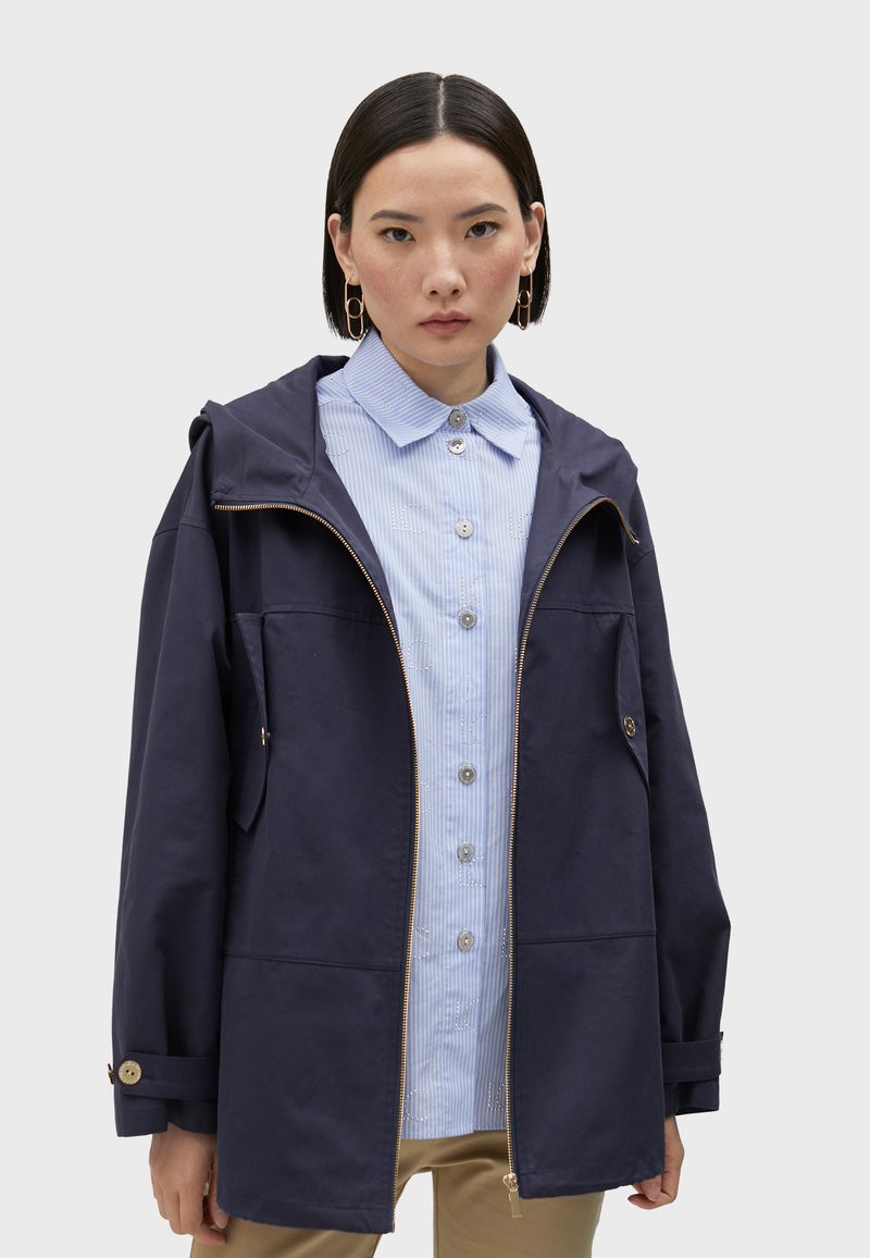 Navy blue hooded jacket with a zip front and side pockets, worn over a light blue striped shirt with round buttons. Cotton blend fabric.