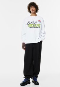 Weißes Langarmhemd mit dem Grafikdesign "STUJD Racing Team" in Grün und Schwarz. Schwarze lockere Hose und schwarze Stiefel mit blauen Schnürsenkeln. Strukturierte Ärmel.