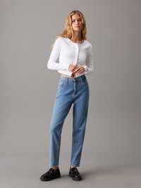 Hosszú szőke hajú nő fehér gombos cropped kardigánt, kék magas derekú farmert és fekete loafert visel, szürke háttér előtt állva.