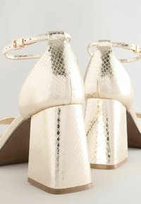 Arany metál magassarkú cipők, kígyóbőr mintázattal. Tömbsarokkal és állítható bokapánttal rendelkeznek.