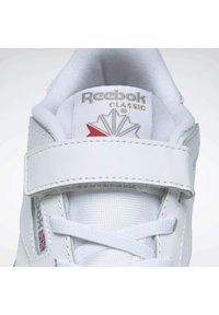 Zapatilla blanca con parte superior de cuero liso, que presenta una cinta de Velcro, cordones y un logo en relieve. Incluye un diseño decorativo en plata y rojo.