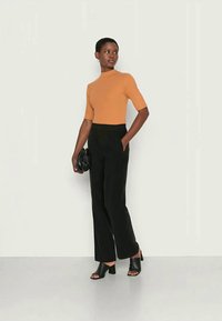 Top laranja justo de mangas curtas, combinado com calças pretas de cintura alta. O modelo segura uma bolsa preta e usa mules pretos de ponta aberta.