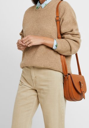 Person iført en beige sweater over en blå blomster skjorte, lyse beige bukser og bærer en brun læder crossbody taske med spænde.