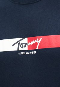 Navy blue fabric with a horizontal white and red flag design, featuring stylized "Tommy" and "JEANS" text below the flag.