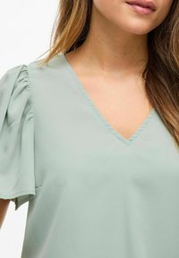 Majica mint zelene boje sa V-izrezom i kratkim, napuhanim rukavima s nabrizanim detaljima. Mekana tekstura s laganom, lepršavom tkaninom.