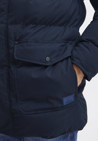Navy blue puffer jacket featuring a zipped front, large snap-button pockets, and a textured patch label on the lower left side.