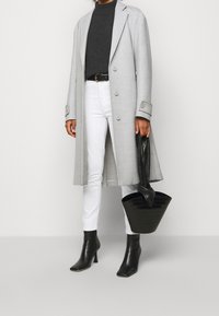 Personne portant un long manteau gris clair, un col roulé noir, un pantalon blanc, des bottes noires à talons, et tenant un sac à main noir avec des poignées torsadées.