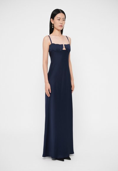 Sieviete tērpusies tumši zilā, līdz grīdai sniedzošā kleitā ar plānām siksnām un strukturētu, zobainu krūšdaļas detalizējumu, valkājot krītošas auskarus.