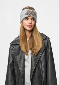 Fascia per capelli grigia lavorata a maglia con finitura testurizzata e decorazione centrale a forma di foglia con perline, indossata da una persona con giacca in pelle nera.