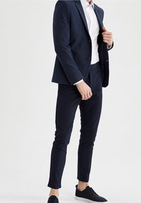 Blazer blu navy con finitura testurizzata, abbinato a una camicia bianca e pantaloni scuri su misura. Indossando sneakers nere, i dettagli includono una chiusura a un solo bottone.