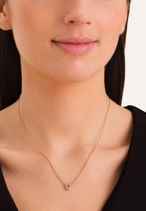 Collana d'oro con una catena sottile e un pendente a forma di U impreziosito da piccoli brillanti, indossata su un collo nudo.