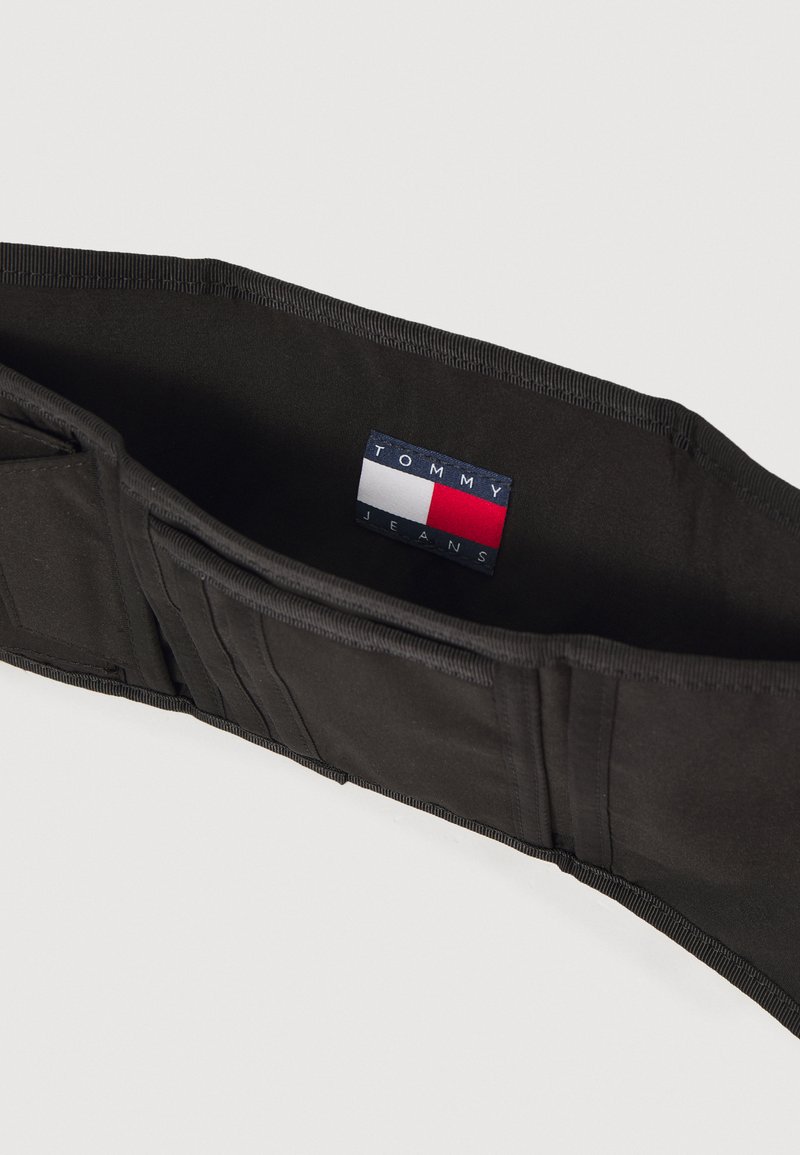 Cinturón elástico negro con múltiples ranuras y una etiqueta de tela con colores rojo, blanco y azul, que dice "TOMMY JEANS."