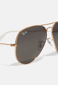 Ochelari de soare Ray-Ban cu un cadru metalic aurit, lentile de un gri închis și pernuțe pentru nas. Prezintă un logo distinctiv pe lentila superioară.