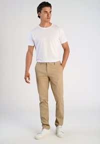 Hombre de pie vistiendo una camiseta blanca sencilla, pantalones beige y zapatillas blancas, con una mano en el bolsillo, mirando hacia su derecha.