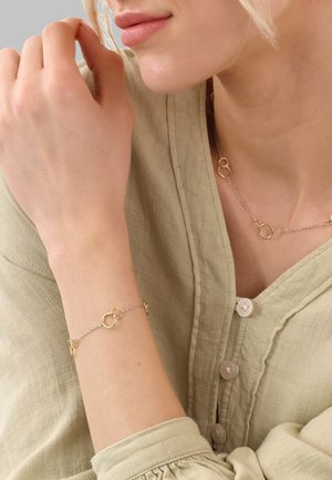 Gold and silver bracelet featuring interlinked circular charms on a delicate chain, worn on a wrist against a textured, light green blouse.