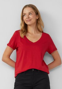 T-shirt rouge à manches courtes avec un col en V, fabriqué en tissu doux. Manches retroussées et coupe décontractée. Associé avec un pantalon noir.
